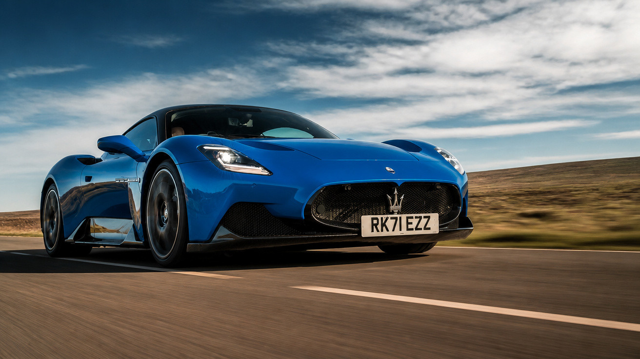 Carro desportivo azul Maserati em movimento numa estrada com céu nublado e paisagem árida ao fundo.