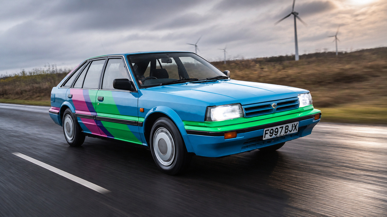 Carro azul com decoração verde e roxa a circular numa estrada molhada sob céu nublado e turbinas eólicas ao fundo.