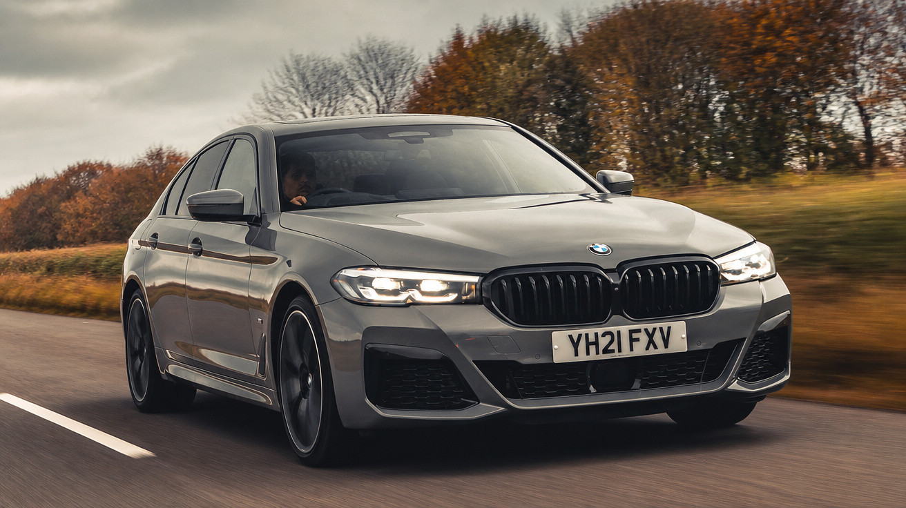 Carro BMW cinzento em movimento numa estrada com árvores e céu nublado ao fundo.