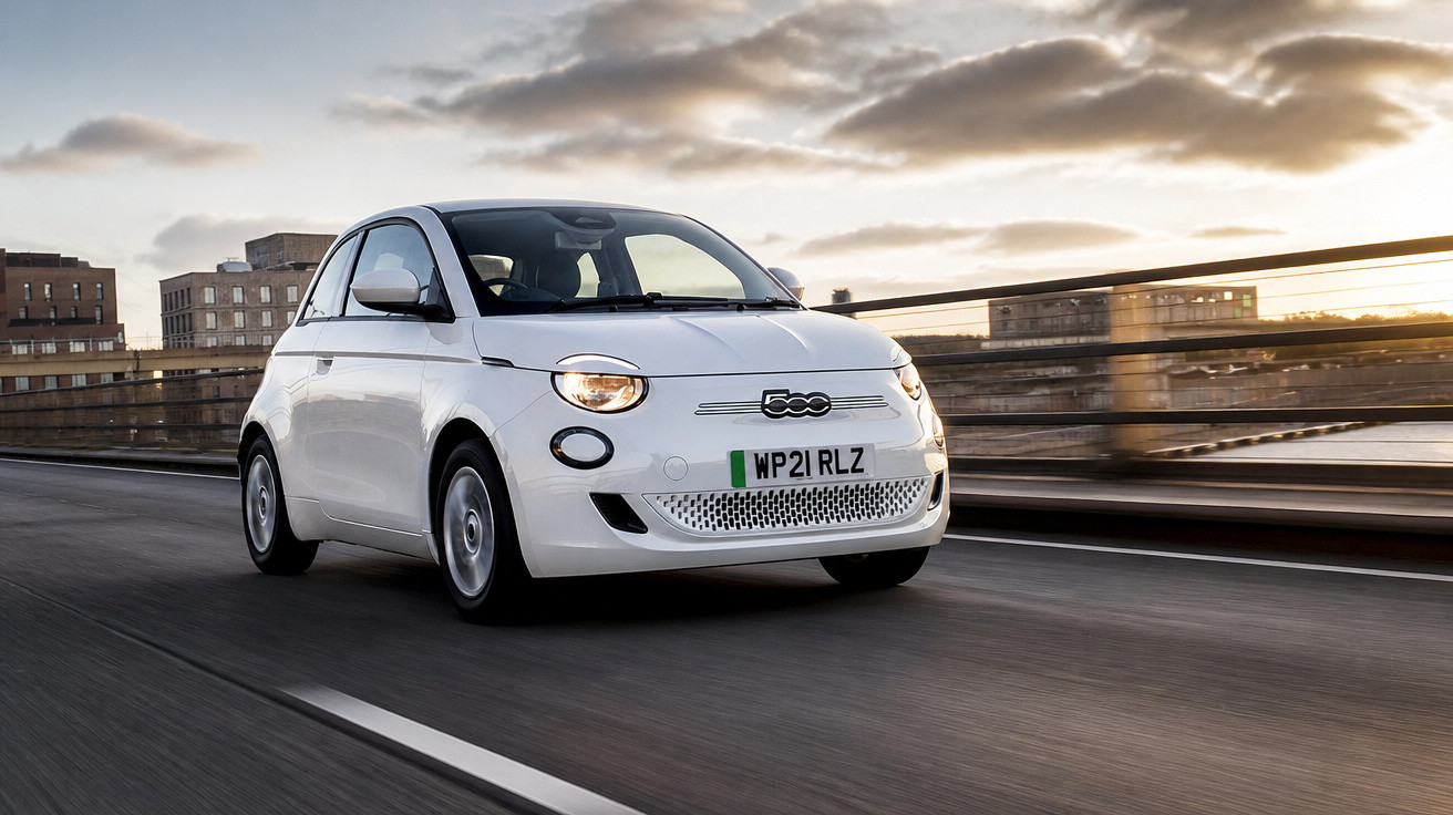 Carro elétrico branco Fiat 500 a circular numa estrada urbana ao pôr do sol.