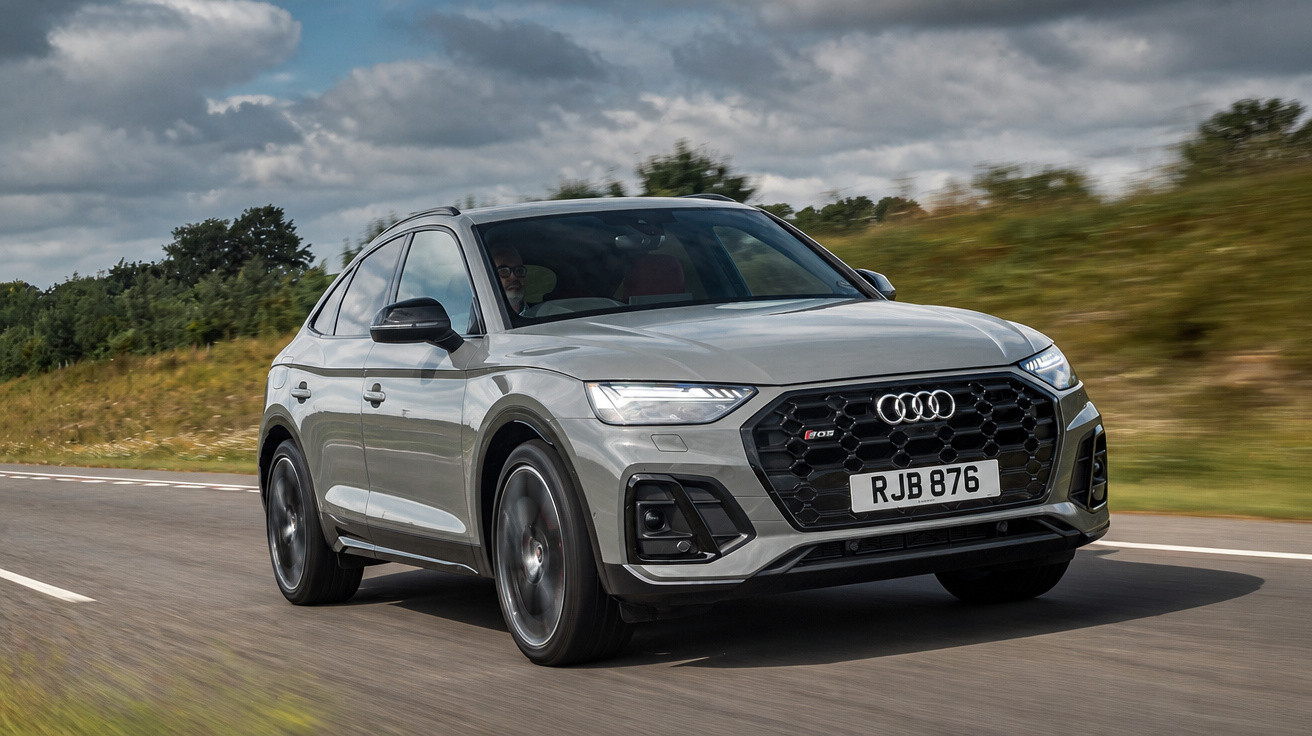 Audi Q5 cinzento moderno a circular numa estrada com vegetação e céu nublado ao fundo.